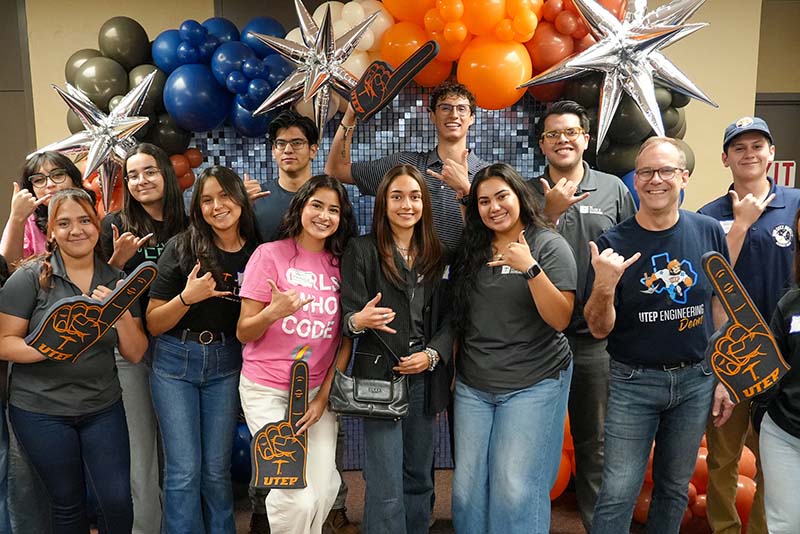 Group of attendees pose together during a UTEP Engineering homecoming gathering while holding a large “Metallurgical, Materials and Biomedical Engineering” banner, seated and standing in front of a balloon-decorated backdrop while making hand gesture signs for the departmental group photo.